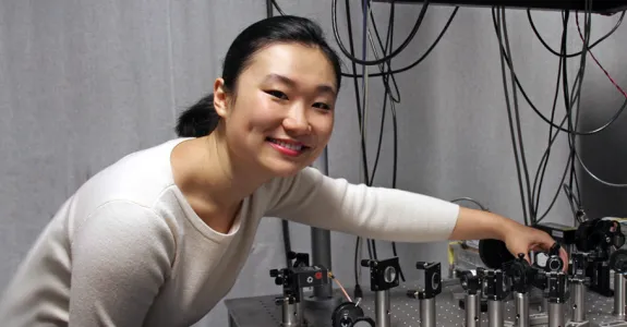 Smiling Asian female graduate student with her hair tied back, wearing a white sweater and leaning down over a complex microscope/laser contraption on a silver table.