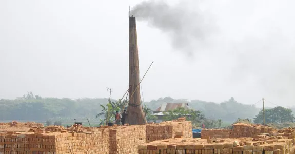 Photo of a tall kiln tower streaming smoke with many huge stacks of orange/red bricks in front of it.