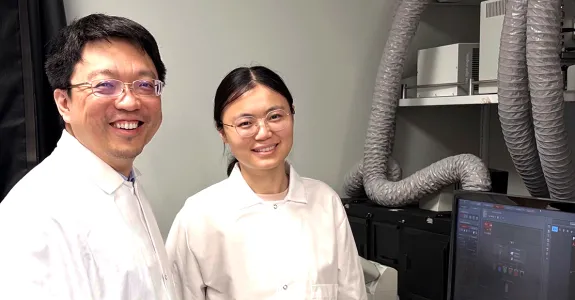 Photo of male faculty member and female postdoc standing in the lab, wearing white lab coats and smiling, with a computer screen showing cell images.