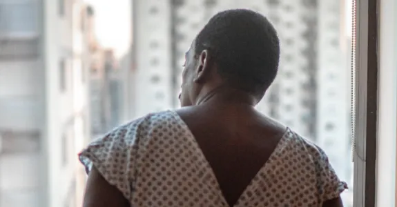 Photo of the back of a man in a hospital gown, in front of a window through which tall buildings are visible.