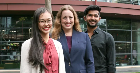 Outdoor photo of a young Asian female medical student, a white female faculty member, and a young southeast Asian male undergraduate student smiling at the camera together.