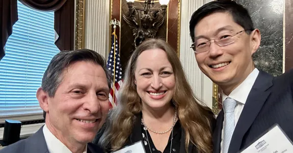 Indoor "selfie" photo of white male faculty member with short gray hair, white female faculty member with long blonde hair, and Asian male faculty member with short black hair and glasses, all smiling at the camera. They are dressed formally, and there are ornate wall decorations showing behind them.