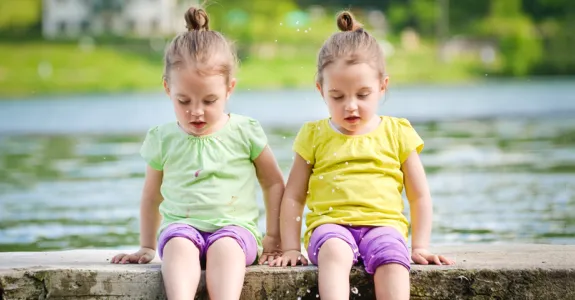 Photo of identical twin toddler-aged girls sitting on a pier with their feet in a lake.