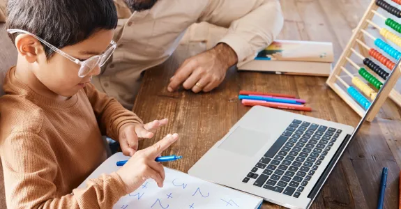 Stock photo of a man sitting with a young child who is counting with their fingers in front of a piece of paper with addition equations.