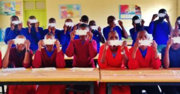 Photo of children in a Ugandan classroom holding up paper Foldscope microscopes.