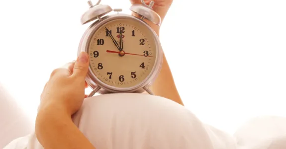 Photo of a pregnant woman lying on her back, holding an old-fashioned alarm clock on top of her stomach.