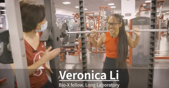 Screenshot from video of an Asian woman asking questions of an Asian female graduate stuent holding a barbell on her shoulders in a gym and smiling. A caption reads "Veronica Li, Bio-X Fellow, Long Laboratory." 