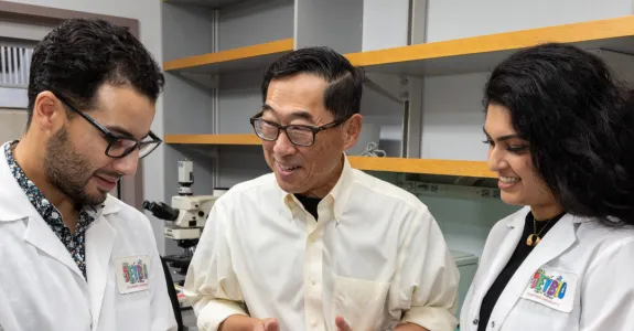 Indoor photo of Stephan Ramos, Professor Seung Kim, and Preksha Bhagchandani in the lab.