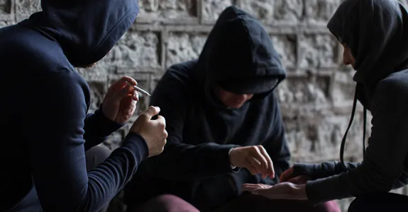 Photo of three adolescents in dark clothes surreptitiously sharing drugs.