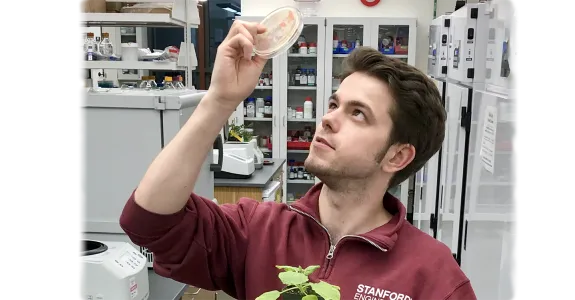 Photo of a male graduate student holding up a petri dish in one hand, with a small plant held in the other.