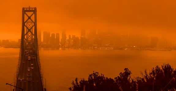 Photo of Bay Bridge in San Francisco, showing the bridge and city skyline wreathed in smoke. Whole photograph is strongly dark orange tinted due to smoke filtering the sunlight.