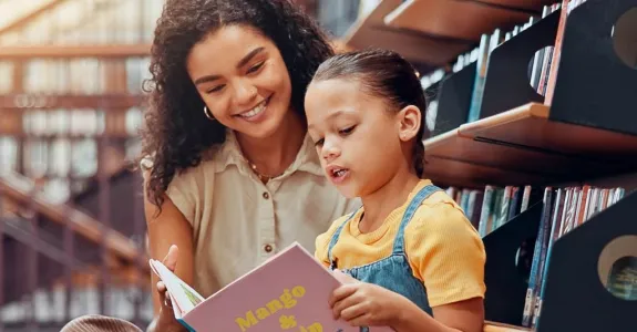 Photo of a child reading from a picture book with a woman looking over her shoulder.