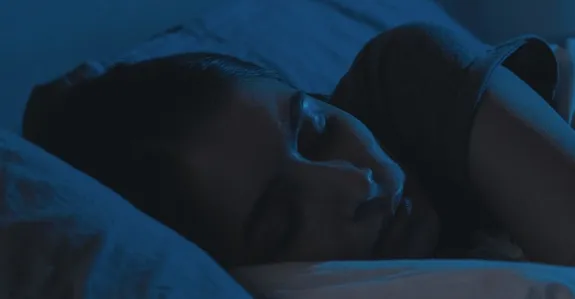 Dark blue tinted photo of a woman's head resting on a pillow as she sleeps.
