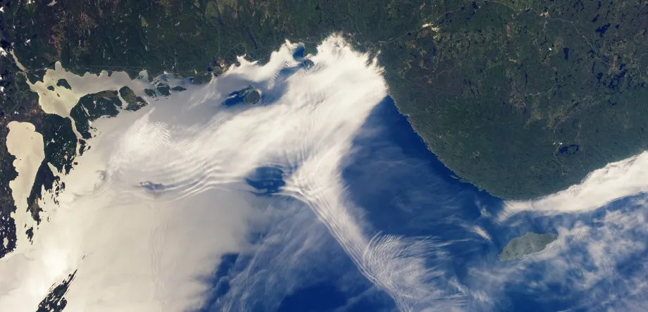 Satellite image of a large body of water and the nearby land, with numerous clouds rippling in different ways.
