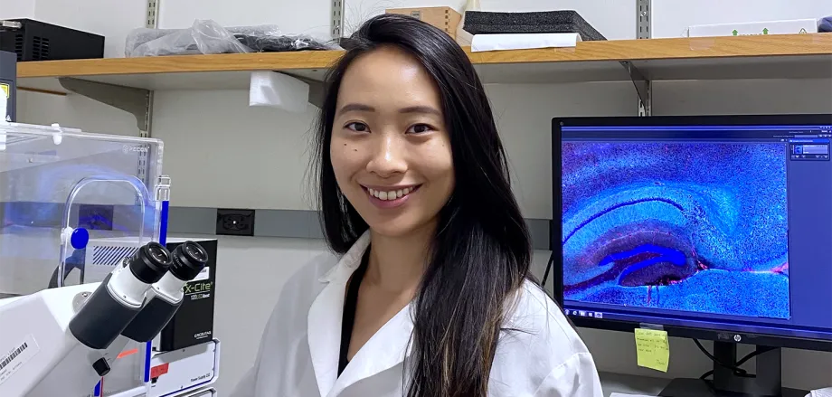 Indoor photo of a smiling Asian female graduate student standing in a wet laboratory next to a microscope, with a computer screen displaying cells in the background.