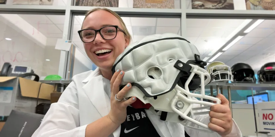 Indoor photo of a white female undergraduate student holding up a football helmet and smiling.