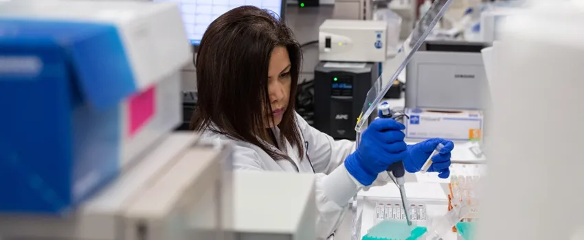 Photo of a female researcher in a lab coat and gloves pipetting into tubes in a laboratory area.