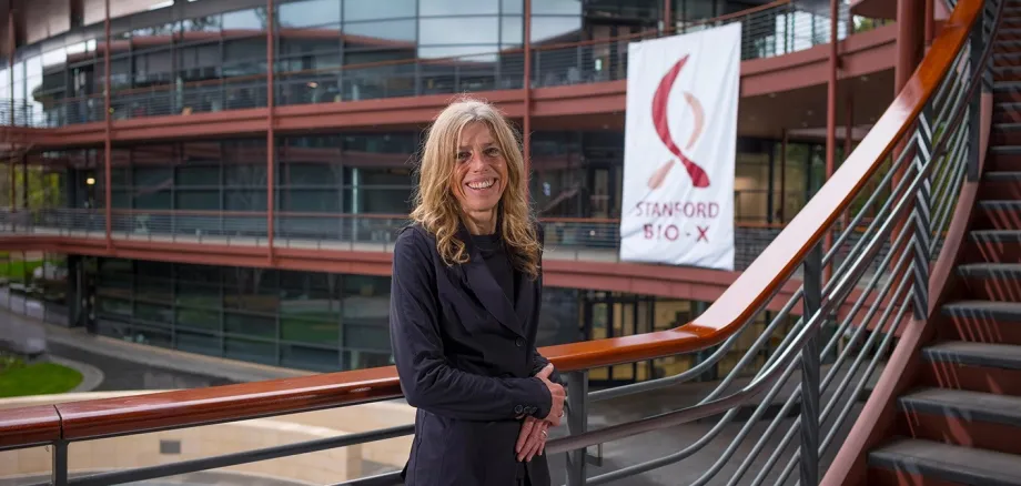 Outdoor photo of a white female faculty member standing on a staircase at the Clark Center, smiling at the camera with a large banner of the Stanford Bio-X logo in the background.