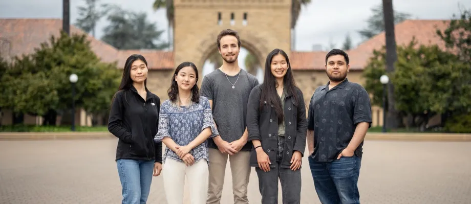 Outdoor photo of 5 undergraduates standing together in front of a Stanford University building.