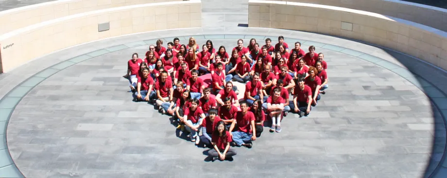 Photo of dozens of Stanford graduate students sitting on the Clark Courtyard outside, wearing matching red shirts. They are seated in a heart shape.