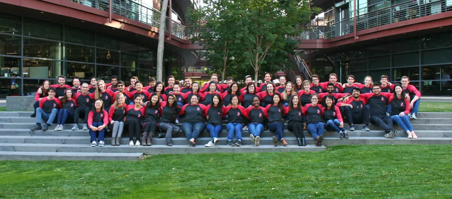 Group photo showing dozens of Stanford Bio-X graduate students seated on steps at the Clark Center, wearing matching shirts and with their arms around each other.