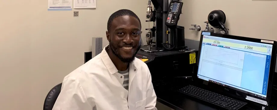 Photo of smiling Black male graduate student sitting in front of a computer screen and a silver and black microscope device, wearing a white lab coat and smiling at the camera.