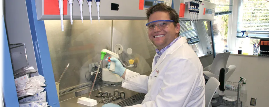 Photo of a smiling male graduate student wearing a white lab coat and other Personal Protection Equipment, sitting at a fume hood and using a pipetting device.