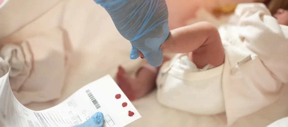Photo of gloved hand holding a small baby's foot and other hand holding a chart with dots of blood.