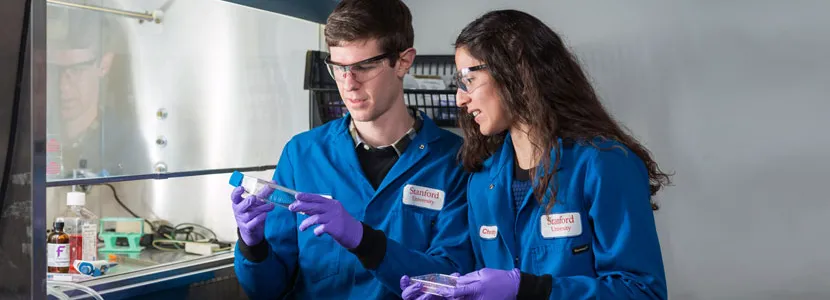 Photo of students Colin McKinlay and Jessica Vargas in the laboratory, examining a test tube and a petri dish near a fume hood.