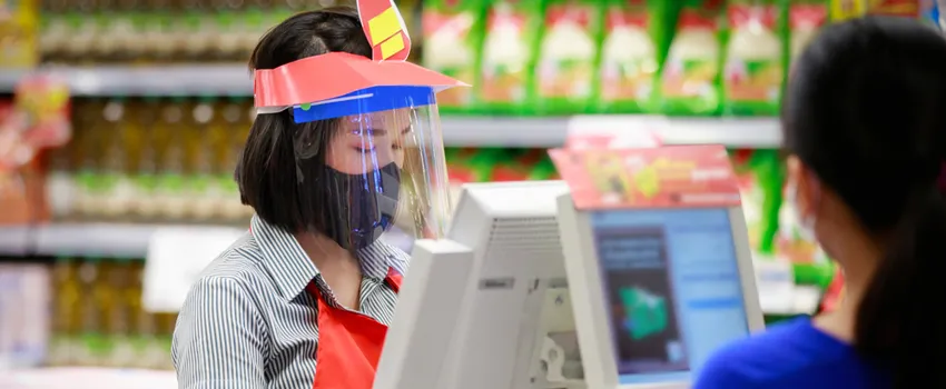 Photo of a female grocery store cashier wearing a mask and a face shield, checking out a shopper wearing a mask.