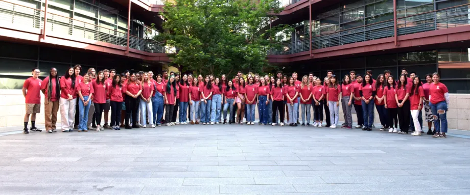 Outdoor photo of 70 Stanford undergraduate students wearing matching pale red T-shirts with the Stanford Bio-X logo and smiling at the camera, all standing in the Clark Center Courtyard.
