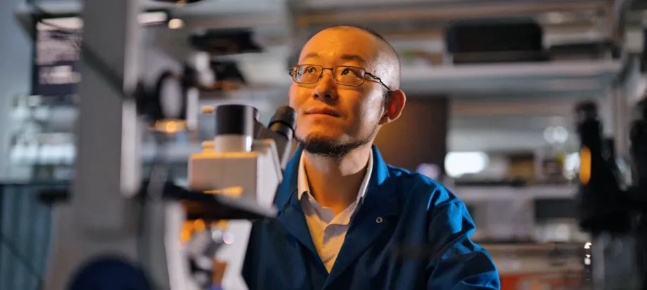 Indoor photo of Dr. Guosong Hong sitting in a laboratory, behind a microscope, looking up and to the left.