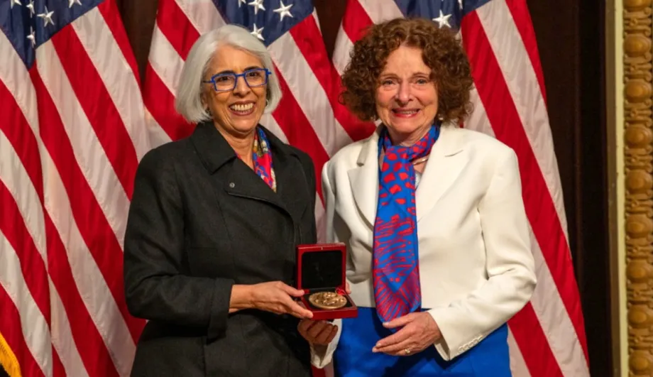 Photo of a southeast Asian woman and a caucasian woman smiling and standing together in front of American flags, holding up a gold medal in a case.