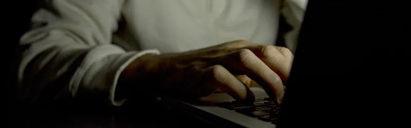 Photo of a man using a computer in the dark, showing only his hand on the keyboard and not his face.