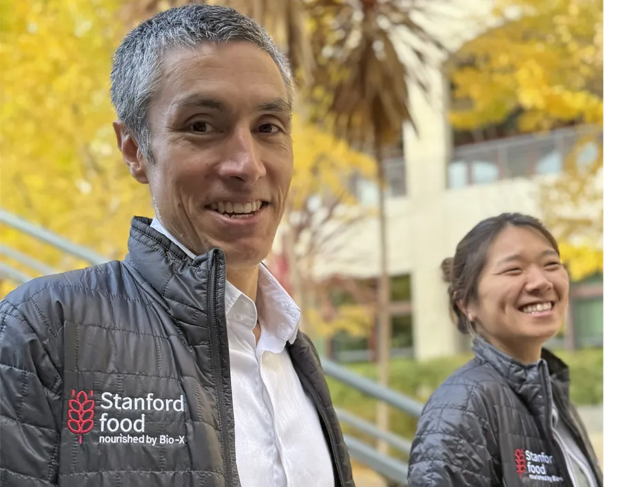 Outdoor photo of Professor KC Huang and postdoc Beverly Fu, both smiling at the camera and wearing Stanford Food branded jackets.
