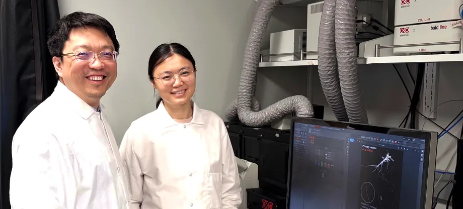 Photo of male faculty member and female postdoc standing in the lab, wearing white lab coats and smiling, with a computer screen showing cell images.