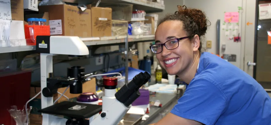 Indoor photo of a Black female graduate student working at a microsocope in a laboratory.