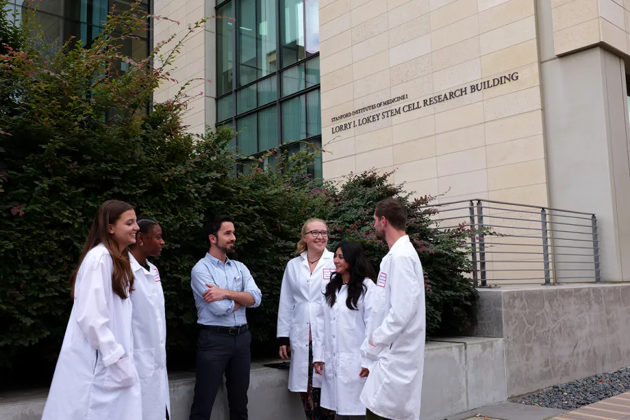 Photo of several researchers standing outside, most of them wearing white lab coats except for male faculty member standing in the middle.