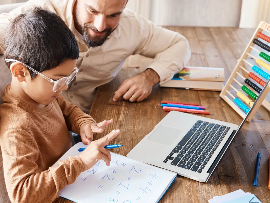 Stock photo of a man sitting with a young child who is counting with their fingers in front of a piece of paper with addition equations.
