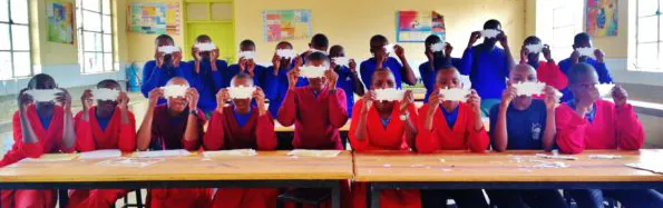 Photo of children in a Ugandan classroom holding up paper Foldscope microscopes.