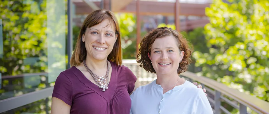 Outdoor photo of two female faculty members standing on a walkway with their arms around each other, smiling.