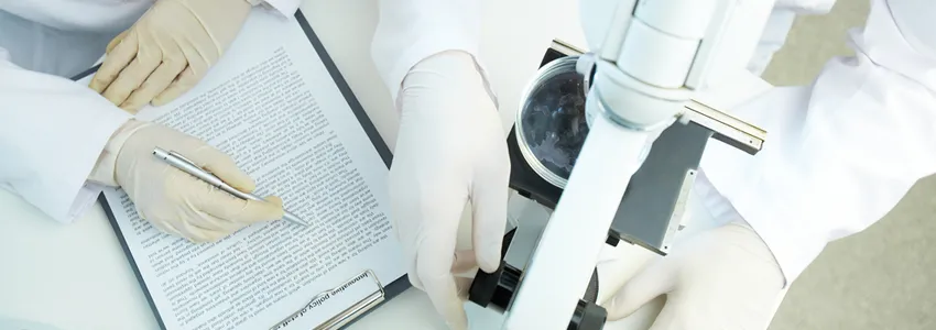 Photo from above of two scientists wearing gloves: one works with a microscope and one has a typed report on a clipboard.