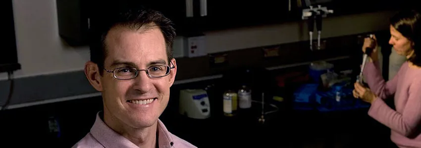 Photo of dark-haired male professor wearing glasses, smiling in lab space with woman pipetting in the background.