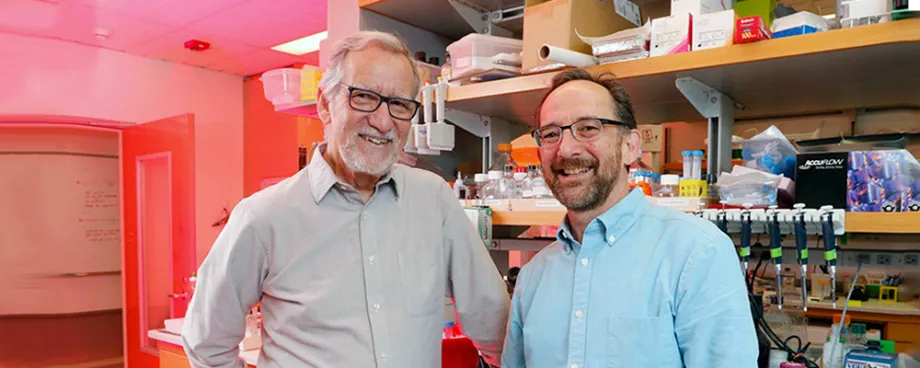 Photo of elder and middle-aged male professors standing in front of shelves with lab equipment and supplies.