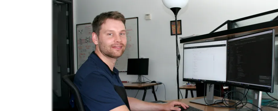 Photo of a white male graduate student wearing a dark blue polo shirt sitting at a beige desk in front of two computer screens. Left screen is white and shows faint lines of code going down the monitor; right screen is black and shows similar lines of code. A whiteboard in the background shows colorful doodles.