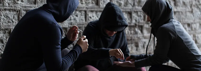 Photo of three adolescents in dark clothes surreptitiously sharing drugs.