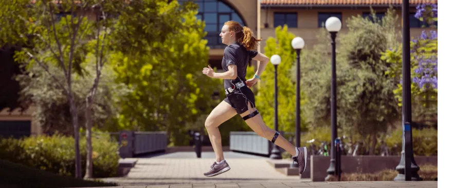 Photo of white female graduate student running across a courtyard area at Stanford, wearing running clothes and the wearable device described in the article.
