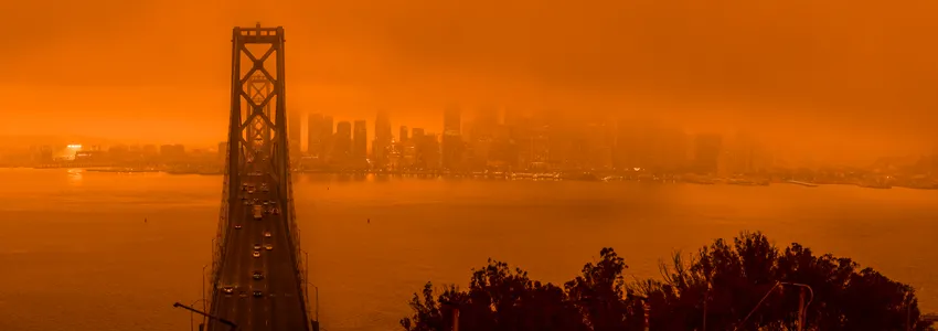 Photo of Bay Bridge in San Francisco, showing the bridge and city skyline wreathed in smoke. Whole photograph is strongly dark orange tinted due to smoke filtering the sunlight.