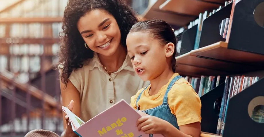 Photo of a child reading from a picture book with a woman looking over her shoulder.