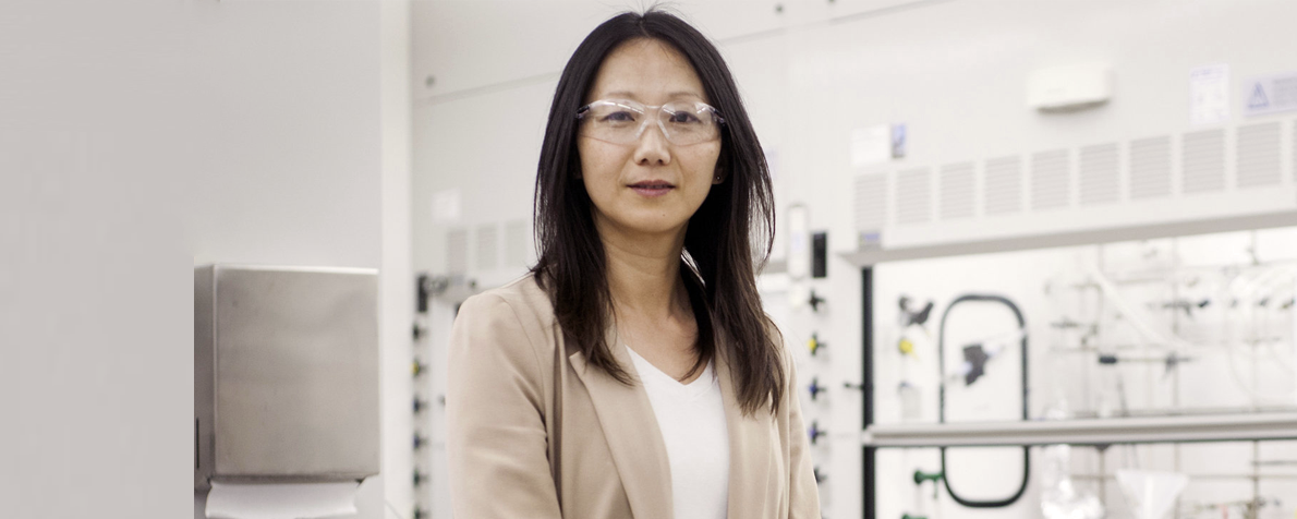 Photo of an Asian female faculty member standing in a very white-toned wet laboratory area, wearing clear protective safety goggles and a beige blazer.